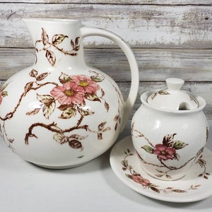 May include: A white ceramic pitcher and sugar bowl set. The pitcher has a large handle and a round body. Both pieces are decorated with pink flowers and brown vines. The sugar bowl sits on a matching saucer.