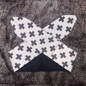 Op de afbeelding: Een zwart-witte haarband met een kruispatroon. De haarband is gemaakt van stof en heeft een zwarte onderkant. De achtergrond is een grijze stof met een spinnenwebontwerp.