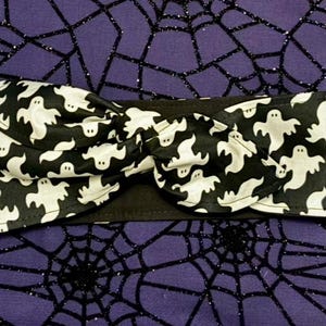 Pode incluir: Faixa de cabeça preta e branca com um padrão de fantasmas. A faixa de cabeça está amarrada em um nó e está em um fundo roxo com um padrão de teia de aranha preta.