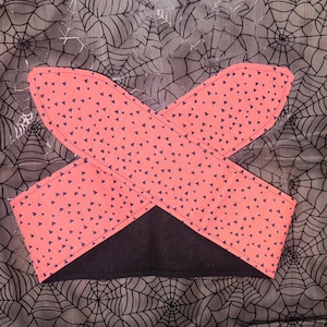 Puede incluir: Una diadema de Halloween con tela de color coral con pequeños triángulos negros. La diadema está cruzada y tiene una base negra. El fondo es una tela gris con un diseño de telaraña.