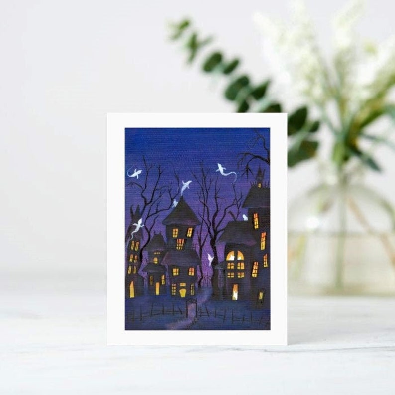 May include: A Halloween-themed greeting card featuring a watercolor painting of a spooky town at night. The houses have lit windows, and ghosts float in the dark blue sky. Bare trees and a picket fence complete the eerie scene.