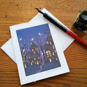 May include: A Halloween-themed greeting card featuring a spooky haunted house scene with ghosts, set against a dark blue sky. A calligraphy pen, ink bottle, and envelope are on a wooden surface.