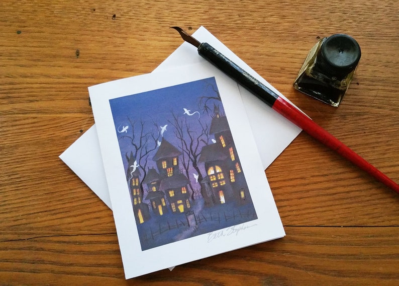 May include: A Halloween-themed greeting card featuring a spooky haunted house scene with ghosts, set against a dark blue sky. A calligraphy pen, ink bottle, and envelope are on a wooden surface.