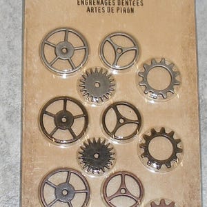 May include: A package of 12 metal sprocket gears in various sizes and designs. The gears are in shades of silver and bronze, and are arranged on a cardboard backing. The package includes the text "idea-ology" and "SPROCKET GEARS".
