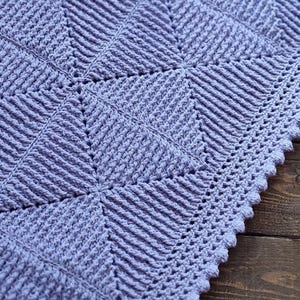 May include: A close-up of a lavender crochet blanket with a geometric pattern. The blanket has a textured design with diagonal lines and a scalloped edge. The blanket is on a wooden surface.