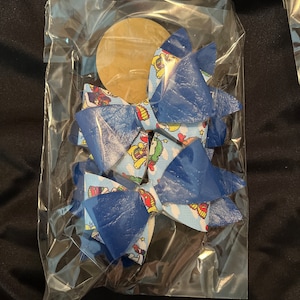 May include: Two hair bows in a clear plastic bag. The bows have a blue leather-like top layer and a patterned fabric underneath with cartoon characters on a light blue background. A round, tan tag is visible.