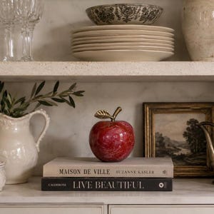 May include: A polished red marble apple sculpture with a metallic gold stem and leaf, resting on a stack of two books titled Maison de Ville by Suzanne Kasler and Live Beautiful by Calderone, positioned on a shelf with other home decor items.