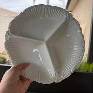 May include: A round white ceramic serving dish divided into three equal sections. The rim features a raised, ornate, scalloped edge with vertical fluted detailing that extends toward the center of the plate, creating a textured surface.