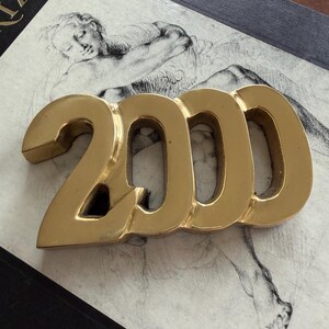 May include: A gold-colored metal sculpture of the number 2000. The numbers are in a bold, sans-serif font. The sculpture is resting on a surface with a sketch of a classical figure.