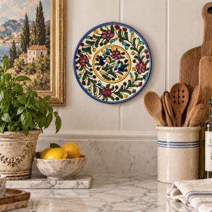 May include: A round ceramic wall plate featuring a hand-painted floral pattern with red flowers, green leaves, and small blue birds on a cream background, framed by a solid blue rim. The design is symmetrical with a central medallion and border.