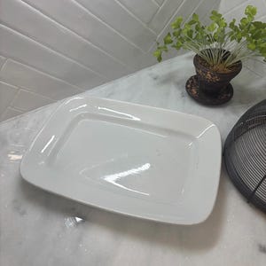 May include: A rectangular white ceramic serving platter with rounded corners and a smooth, glossy finish. The platter features a slightly recessed center area and a wide, flat rim, presenting a clean and simple design for food presentation.
