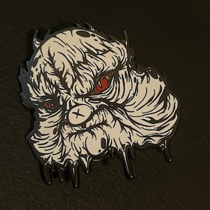May include: A white and black enamel pin featuring a stylized monster face with red eyes. The monster has a cross mark on its face and dripping details. The pin is set against a dark background.