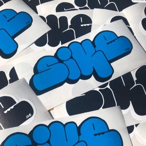 May include: A collection of white stickers with a blue and black graphic design. The stickers feature the word "SIKE" in a bold, bubble-style font, with a blue outline and a black fill. Some stickers have a black graphic of a face.