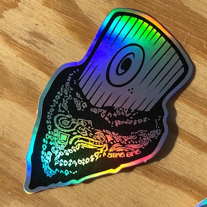 May include: A holographic sticker with a black and white design. The design features a large eye with a circular pupil and a detailed pattern of lines and swirls. The sticker has a rainbow iridescent finish.