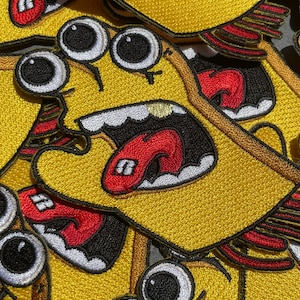 Puede incluir: Un conjunto de parches bordados amarillos con un personaje de dibujos animados con tres ojos, una boca abierta de par en par y una lengua roja. El personaje lleva una camisa amarilla y tiene un diseño rojo y negro en su camisa.