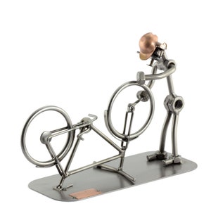 Könnte beinhalten: Eine Metallsculptur einer Person, die ein Fahrrad repariert. Die Skulptur besteht aus Muttern, Schrauben und anderen Metallteilen. Die Person trägt einen Helm und hält einen Schraubenschlüssel. Das Fahrrad steht auf einem Metallsockel.