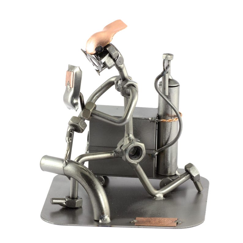 May include: A metal figurine of a welder wearing a welding mask and holding a welding torch. The figurine is made of nuts, bolts, and other metal pieces. The welder is standing on a metal base with a copper plate that reads "Welder".