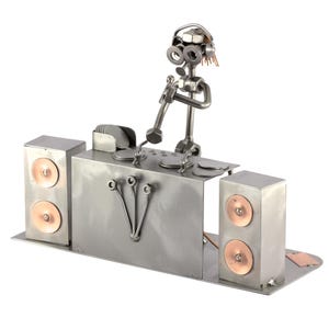 Op de afbeelding: Een metalen sculptuur van een DJ met een koptelefoon, een microfoon en een mengpaneel. De DJ staat op een platform met twee luidsprekers aan weerszijden. Het beeld is gemaakt van verschillende metalen onderdelen, waaronder moeren, bouten en ringen.