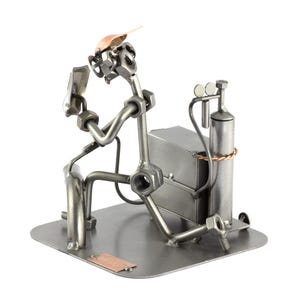 May include: A metal figurine of a welder wearing a welding mask and holding a welding torch. The figurine is made of nuts, bolts, and other metal pieces. The welder is standing next to a welding tank and a welding table.