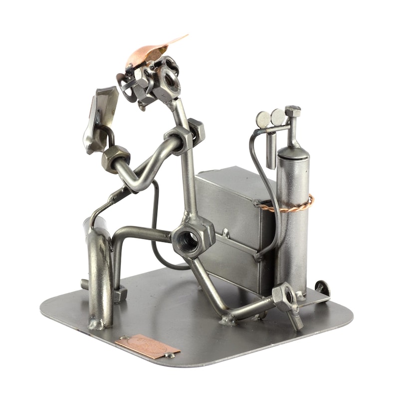 May include: A metal figurine of a welder wearing a welding mask and holding a welding torch. The figurine is made of nuts, bolts, and other metal pieces. The welder is standing next to a welding tank and a welding table.