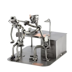 Puede incluir: Una escultura de metal de dos figuras, una sentada en un escritorio y la otra en una silla. Las figuras están hechas de tuercas, tornillos y otras piezas de metal. El escritorio tiene una lámpara y un cajón. La escultura es una representación caprichosa de un médico examinando a un paciente.