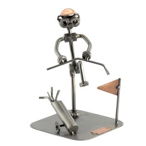 May include: A metal sculpture of a golfer in a golf stance, holding a golf club. The sculpture is made of nuts, bolts, and other metal pieces. The golfer is wearing a copper-colored hat. The sculpture is on a metal base with a golf bag and a flag.