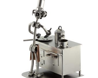 May include: A metal figurine of a chef cooking on a stove. The chef is made of nuts, bolts, and other metal pieces. The stove is also made of metal and has a pot and a pan on it.
