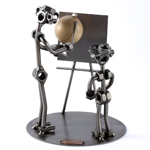 Könnte beinhalten: Eine Metallskulptur aus zwei Figuren, die aus Muttern und Schrauben bestehen. Die Figur links hält einen Globus aus Holz und zeigt auf eine Tafel, die von der Figur rechts gehalten wird.