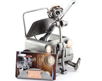 May include: A metal sculpture of a mechanic holding a wrench, standing next to a car with the hood open. The sculpture is made of nuts, bolts, and other metal pieces. The sculpture is holding a business card with the company name "Steelman24" and contact information.