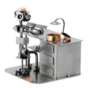 Pode incluir: Uma figurinha de metal de uma pessoa sentada em uma mesa com uma lâmpada, ferramentas e uma pequena unidade de gavetas. A figurinha é feita de porcas, parafusos e outras peças de metal.