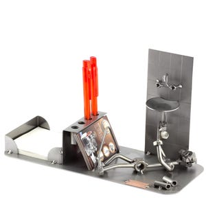 Puede incluir: Organizador de escritorio de metal con una figura de fontanero, un porta bloc de notas y un porta bolígrafos. La figura es de metal y sostiene una llave inglesa. El porta bloc de notas es de metal y tiene un bloc de notas blanco. El porta bolígrafos es de metal y tiene tres bolígrafos rojos.