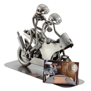 May include: A metal sculpture of two figures riding a motorcycle. The figures are made of nuts, bolts, and other metal pieces. The motorcycle is also made of metal and has a detailed design. The sculpture is on a metal base with a business card holder.
