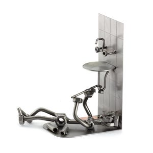 Puede incluir: Una escultura de metal de un fontanero acostado de espaldas, rodeado de herramientas, con un lavabo y un grifo en la pared detrás de él.