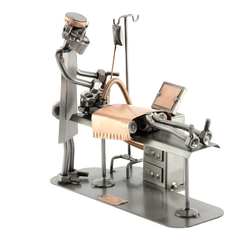 May include: A handcrafted metal sculpture depicting a surgical scene. The sculpture features a doctor figure, operating table, and medical equipment. The doctor is wearing a mask and gown, and the patient is on the table. The sculpture is made of silver and copper-colored metal.