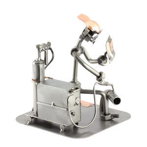May include: A metal figurine of a welder working on a welding machine. The figurine is made of various metal pieces, including nuts, bolts, and screws. The welder is wearing a welding mask and holding a welding torch. The welding machine is on wheels and has a tank on the side.