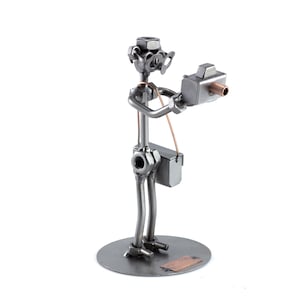 Peut inclure: Une figurine en métal d'un photographe tenant un appareil photo. La figurine est faite de noix, de boulons et d'autres pièces métalliques. Le photographe porte un chapeau et des lunettes. La figurine est debout sur une base en métal.