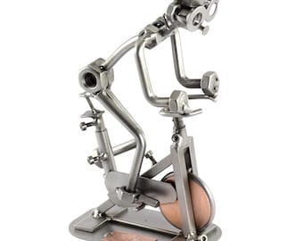May include: A silver metal sculpture of a person riding a stationary bike. The sculpture is made of nuts, bolts, and other metal pieces. The bike has a copper-colored wheel.