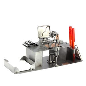 Könnte beinhalten: Ein Schreibtisch-Organizer aus Metall mit einer Figur eines Menschen, der an einem Schreibtisch mit einem Computer sitzt. Der Schreibtisch hat einen Stifthalter, einen Notizblockhalter und ein kleines Regal. Die Figur ist aus Metall und silberfarben.