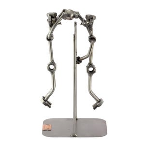 Op de afbeelding: Een metalen sculptuur van een persoon die volleybal speelt. De sculptuur is gemaakt van moeren, bouten en metalen staven. De persoon is in de lucht en slaat de bal. De sculptuur staat op een metalen voet.