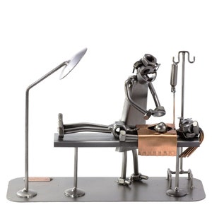 Puede incluir: Una escultura de metal de un cirujano operando a un paciente. El cirujano está hecho de tuercas, tornillos y otras piezas de metal. El paciente está acostado en una mesa con una lámina de cobre que cubre su pecho. La escultura está sobre una base con una lámpara.