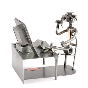 Puede incluir: Una escultura de metal de una persona sentada en un escritorio con una computadora. La escultura está hecha de tuercas, tornillos y otras piezas de metal. La persona lleva gafas y corbata. El escritorio tiene un cajón y un estante.