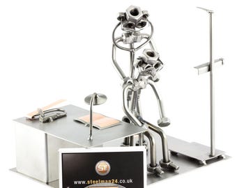 May include: A metal sculpture of two figures sitting at a desk with a lamp and a book. The sculpture is made of nuts, bolts, and other metal pieces. The sculpture is a business card holder and has a business card with the website www.steelman24.co.uk on it.