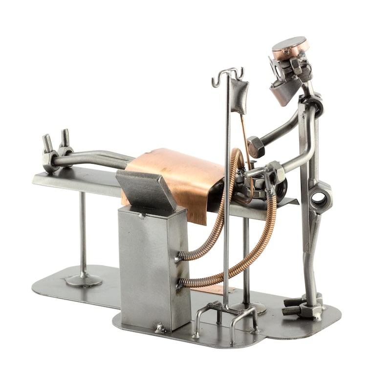 May include: A metal sculpture depicting a surgical scene. A patient lies on a table covered with copper sheeting. A doctor figure, constructed from metal nuts and bolts, stands over the patient. An IV drip and other medical equipment are also present.
