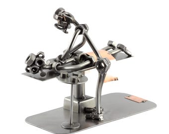 Può includere: Scultura in metallo raffigurante un dentista e un paziente. Le figure sono costruite con dadi, bulloni e aste metalliche, con un elemento color rame. La scultura è montata su una base metallica rettangolare.
