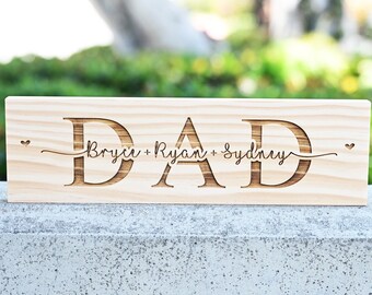 May include: Wooden sign with the word "DAD" in large, carved letters. The names "Bryce", "Ryan", and "Sydney" are written in a smaller font below the word "DAD" with hearts on either side.