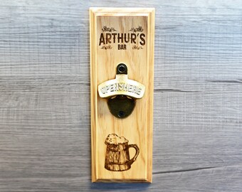 May include: A wooden wall mounted bottle opener with the text "Arthur's Bar" and "Open Here" engraved on it. The bottle opener has a metal opener and a woodburned image of a beer stein.