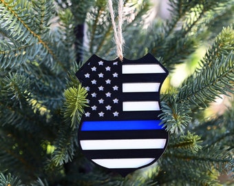 May include: A wooden Christmas ornament shaped like a police badge with an American flag design and a blue line across the bottom.