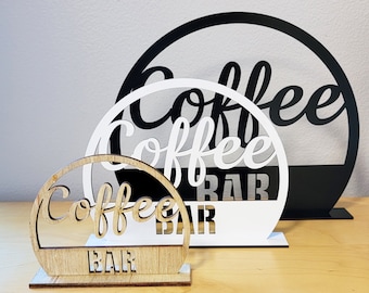 May include: Three wooden signs with the text "Coffee Bar" in black and white lettering. The signs are stacked on top of each other, with the largest sign at the bottom and the smallest sign at the top.