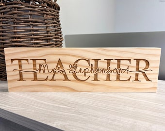 May include: A rectangular wooden sign with the word "TEACHER" and the name "Mrs. Stephenson" engraved in a light brown color. The sign is made of light-colored wood and is likely a desk accessory.