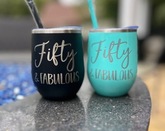 May include: Two stainless steel tumblers with black and turquoise blue finishes. Both tumblers have the text "Fifty & Fabulous" printed on them in silver lettering.